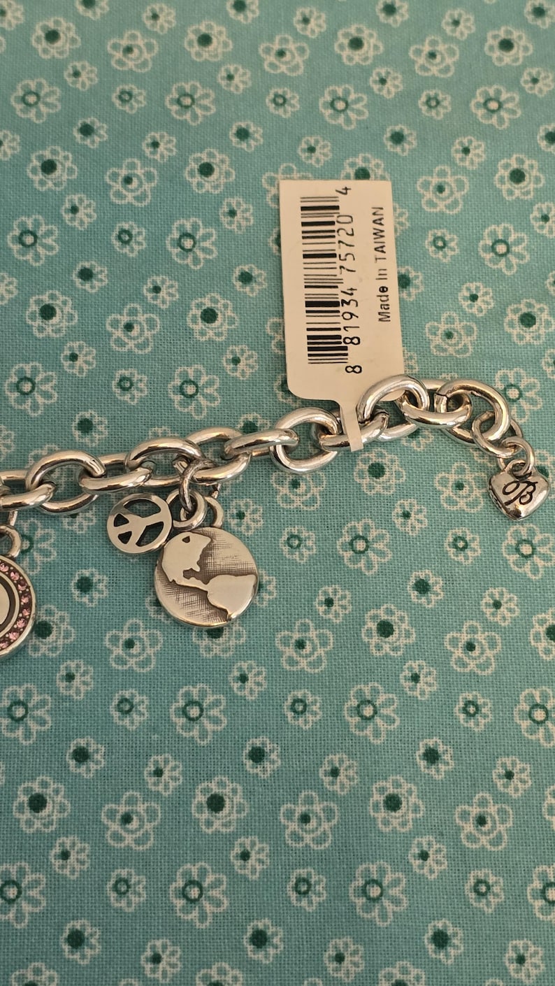 May include: Silver-toned charm bracelet with multiple charms, including a peace sign, a globe, and a heart-shaped charm. The bracelet is displayed on a teal fabric with a floral pattern. A tag with a barcode is visible.