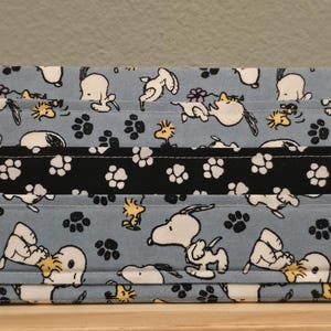 May include: A rectangular fabric pouch featuring a light blue background with a repeating pattern of Snoopy and Woodstock characters, small purple flowers, and black paw prints, accented by a horizontal black band with white paw prints.