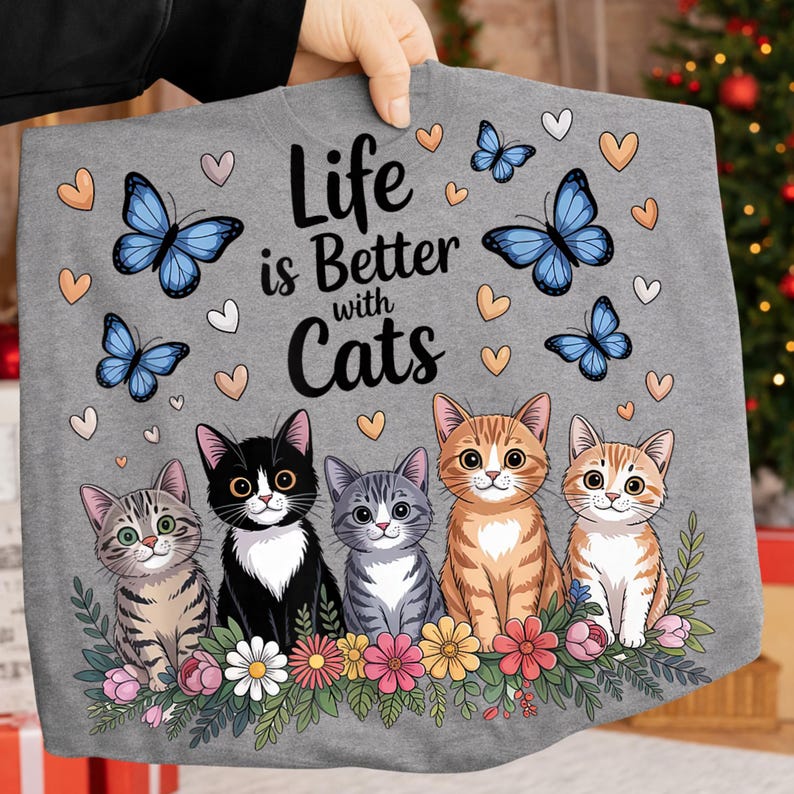 May include: A gray sweatshirt featuring the phrase "Life is Better with Cats" with illustrations of five cartoon cats, blue butterflies, and hearts. The cats are in various colors and patterns, with a floral border at the bottom.