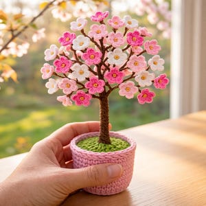 May include: A handcrafted crochet tree with pink, white, and light pink flowers. The tree is in a pink crochet pot with green 'grass'. The trunk is brown. A decorative, miniature, floral accent piece.