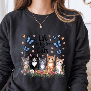 May include: Dark gray sweatshirt with the phrase "Life is Better with Cats" and illustrations of five cats, butterflies, and hearts. The cats are in various colors and patterns, sitting in front of a floral arrangement.