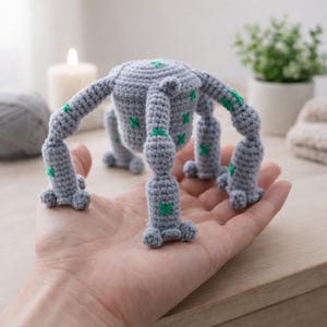 Könnte beinhalten: Ein handgefertigtes graues Häkeltier mit sechs Beinen und einem abgerundeten Körper. Die Beine und der Körper sind mit grünen Kreuzstichdetails verziert. Das Spielzeug wird in der Hand einer Person gehalten, um seine Größe und Handwerkskunst zu zeigen.