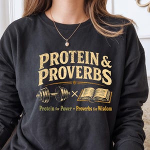 Puede incluir: Sudadera negra con el texto "PROTEIN & PROVERBS" en fuente dorada desgastada. Debajo, una pesa cruzada con un libro abierto. El texto "Protein for Power • Proverbs for Wisdom" está debajo.