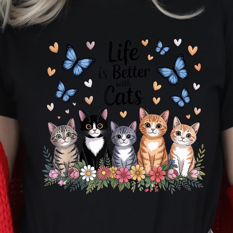 May include: Black t-shirt with a graphic featuring five cartoon cats, butterflies, and hearts. The text "Life is Better with Cats" is displayed above the cats. The cats are in various colors and patterns.