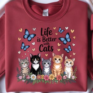 May include: Burgundy sweatshirt featuring a graphic of five cartoon cats, butterflies, and hearts. The text "Life is Better with Cats" is displayed above the cats. The cats are sitting on a bed of flowers.