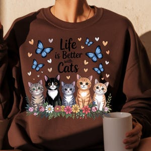 May include: Brown sweatshirt with the phrase "Life is Better with Cats" and an illustration of five cats, butterflies, and hearts. The cats are sitting on a bed of flowers.