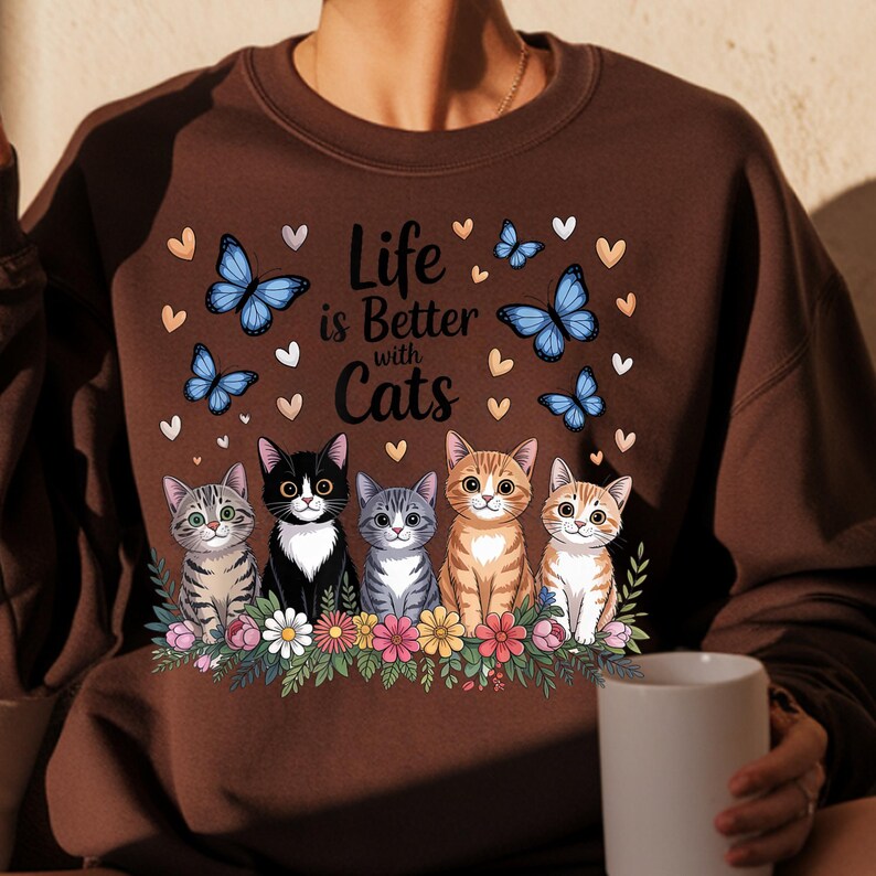 May include: Brown sweatshirt with the phrase "Life is Better with Cats" and an illustration of five cats, butterflies, and hearts. The cats are sitting on a bed of flowers.