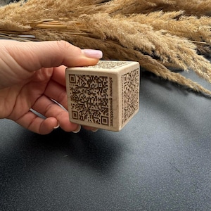 May include: A light brown wooden cube featuring QR codes on all sides. The cube is held in a hand and has the text "закордонний паспорт" printed on one side. The cube is a square shape.