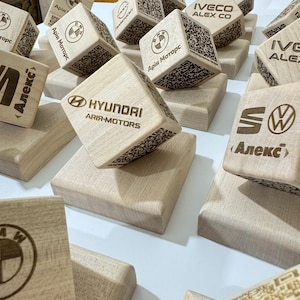May include: Wooden cubes with engraved logos of various car brands, including BMW, Hyundai, and Volkswagen. The cubes are light brown and rest on matching wooden platforms. Some cubes feature QR codes.