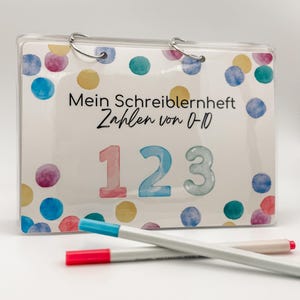 Può includere: Uno strumento didattico con i numeri 1, 2 e 3 in colori pastello. La copertina riporta la scritta "Mein Schreiblernheft Zahlen von 0-10" ed è decorata con pois colorati. Due penne con punte rosse e blu sono in primo piano.