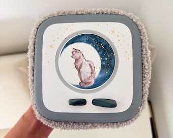 Puede incluir: Una máscara para dormir cuadrada, gris y blanca, con una ilustración de acuarela de un gato en una luna creciente. La máscara tiene un borde suave y difuso y dos botones ovalados de color verde oscuro.