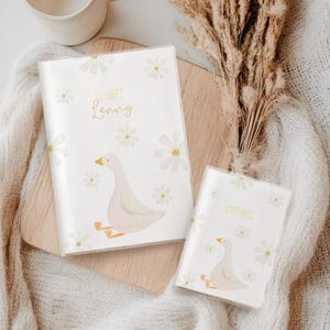 Könnte beinhalten: Zwei weiße Passhüllen mit Gänseblümchen- und Gänsemotiv. Auf einer Hülle steht "JI-HEFT Lenny" in goldener Schrift, auf der anderen "IMPFPASS". Die Gans ist hellbeige mit gelben Füßen und Schnabel. Der Hintergrund ist weiß mit verstreuten Gänseblümchen.
