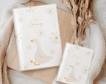 Könnte beinhalten: Zwei weiße Passhüllen mit Gänseblümchen- und Gänsemotiv. Auf einer Hülle steht "JI-HEFT Lenny" in goldener Schrift, auf der anderen "IMPFPASS". Die Gans ist hellbeige mit gelben Füßen und Schnabel. Der Hintergrund ist weiß mit verstreuten Gänseblümchen.