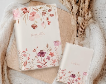Könnte beinhalten: Zwei cremefarbene Notizbücher mit floralen Aquarelldesigns. Das größere Notizbuch trägt den Namen "Mariella" und das Datum "08.08.2023". Das kleinere Notizbuch trägt das Wort "Impass". Das florale Design umfasst rosa und bordeauxfarbene Blumen.