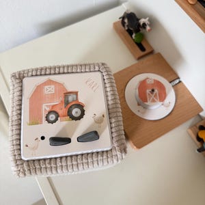 Könnte beinhalten: Ein beige gerahmtes Spielzeug mit einer Bauernhofszene mit einem roten Traktor, einer Scheune und Hühnern. Der Name "KARL" ist oben rechts aufgedruckt. Zwei graue Knöpfe befinden sich darunter. Ein Holzspielzeug mit einer Scheune ist ebenfalls sichtbar.