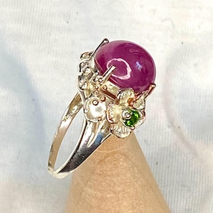May include: A sterling silver ring featuring a large, oval-shaped, deep pink gemstone. The ring has a floral design with a small green stone accent. The ring is displayed on a wooden stand.
