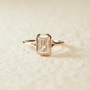 Pode incluir: Um anel de ouro com um diamante lapidação esmeralda. O diamante é engastado em uma moldura e a banda é de ouro polido simples. O anel é exibido em uma superfície branca.