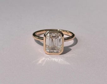 May include: A gold ring with a large, rectangular, clear gemstone. The gemstone is set in a bezel setting, and the band is a simple, polished gold. The ring is displayed on a white surface.