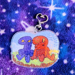 Peut inclure: Un porte-clés en forme de cœur avec deux créatures de dessin animé, une violette et une rouge, assises sur de l'herbe. Le porte-clés a un fermoir argenté et une finition irisée scintillante.