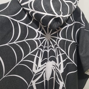 Può includere: Felpa con cappuccio grigio scuro con un grande disegno a ragnatela bianca sul retro e sul cappuccio. Un ragno bianco è centrato all'interno della ragnatela. La felpa ha un cappuccio con coulisse e una tasca a marsupio.