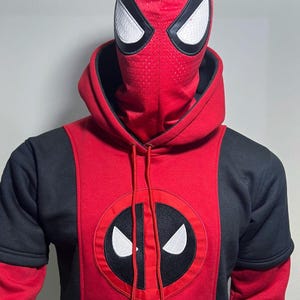 May include: A red and black hooded sweatshirt featuring a central circular emblem with a stylized mask graphic. The hood includes an integrated red mesh face covering with white eye patches. The text KUSTOMLEATHERDEN is printed at the bottom.