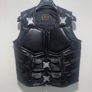 May include: A black leather vest featuring padded chest panels, four front pockets with flaps, and silver-toned metal quick-release buckles on the shoulders and front closure. The interior label displays the text KLD in red lettering.