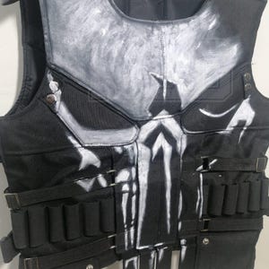 Könnte beinhalten: Schwarze taktische Weste mit weißem Totenkopf-Design auf der Vorderseite. Die Weste hat mehrere Taschen, Gurte und einen gepolsterten Schulterbereich. Die Weste ist zum Tragen über der Kleidung konzipiert.