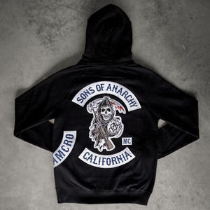 Puede incluir: Sudadera con capucha negra con el logo de Sons of Anarchy en la espalda. El diseño incluye una parca con una guadaña y una pistola, junto con las palabras "Sons of Anarchy" y "California". La sudadera también tiene la palabra "MCRO" en la manga izquierda.