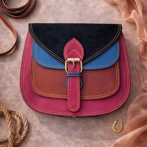 Könnte beinhalten: Eine mehrfarbige Leder-Umhängetasche mit Schnallenverschluss. Die Tasche hat eine schwarze Klappe, blaue und rote Einsätze und eine rosa Basis. Ein goldener Hufeisen-Anhänger und ein Seil sind ebenfalls sichtbar.