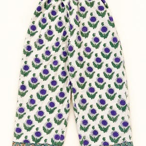 May include: Wide-leg trousers featuring an elasticated waistband and a repeating floral pattern of purple flower heads with green leaves on a white background. The hem is finished with a contrasting band featuring a smaller, repeating green and tan leaf print.