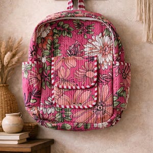 Puede incluir: Mochila rosa con estampado floral y ribete a rayas blancas y rojas. La mochila tiene un gran estampado floral con detalles en blanco y verde. Cuenta con dos bolsillos laterales y un bolsillo frontal con borde a rayas. Cuelga de un gancho de madera.