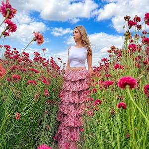 Peut inclure: Un haut court sans manches blanc associé à une longue jupe maxi à étages ornée d'un motif floral rose et rouge. La jupe présente plusieurs couches horizontales à volants qui créent une silhouette volumineuse descendant jusqu'au sol.