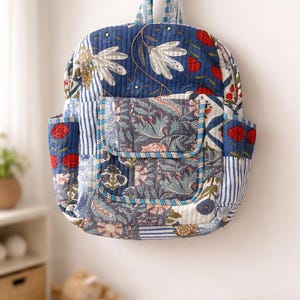 May include: Quilted backpack with a patchwork design. The bag features floral patterns in blue, red, and white. It has a top handle and two side pockets. The backpack is hanging on a wooden peg.