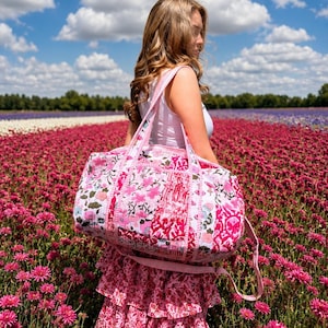 May include: A large, rectangular quilted duffel bag featuring a vibrant pink and white floral block-print pattern. The bag has two sturdy fabric handles and a matching shoulder strap, with visible stitching throughout the quilted fabric construction.