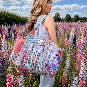 Può includere: Una borsa da viaggio trapuntata con un design patchwork di vari motivi floreali e a righe nelle tonalità del blu, rosa, rosso e bianco. La borsa presenta due tracolle e una forma rettangolare con cuciture visibili su tutto il tessuto.