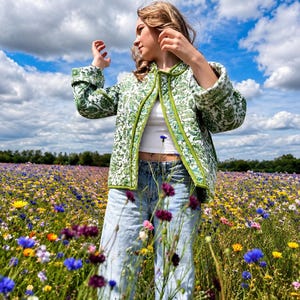 May include: A quilted jacket featuring a green and white floral pattern with solid green trim, worn over a white tank top and light blue denim jeans. The jacket has long sleeves with folded cuffs and a straight hemline, showcasing a detailed texture.