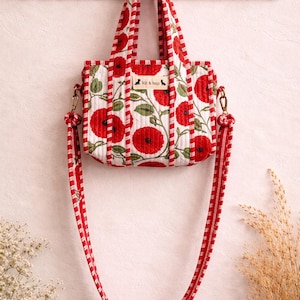 May include: A small, quilted tote bag featuring a red poppy floral design on a white backdrop. It has a matching red and white striped strap and handles. The brand name "kit & hop" is printed on the front.