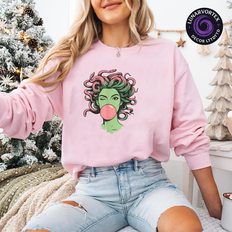 May include: A light pink sweatshirt featuring a graphic of Medusa winking and blowing a bubblegum bubble. The Medusa illustration has green skin, pink and green hair, and is outlined in black. The sweatshirt has long sleeves and a crew neck.