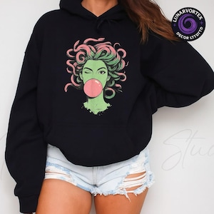 May include: Black hoodie featuring a graphic of a green-skinned woman with pink snakes for hair, winking and blowing a pink bubble. The hoodie has a drawstring hood and a kangaroo pocket. The text "LUNARVORTEX DECOR LTDTTD" is printed on the upper right.