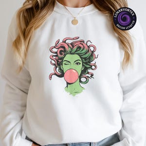 May include: White sweatshirt featuring a graphic of Medusa winking and blowing a bubblegum bubble. The Medusa illustration has green skin, green and pink hair, and is winking. The sweatshirt has a crew neck and long sleeves. The brand name "LUNARVORTEX DECOR LTDTTD" is printed on the upper right.