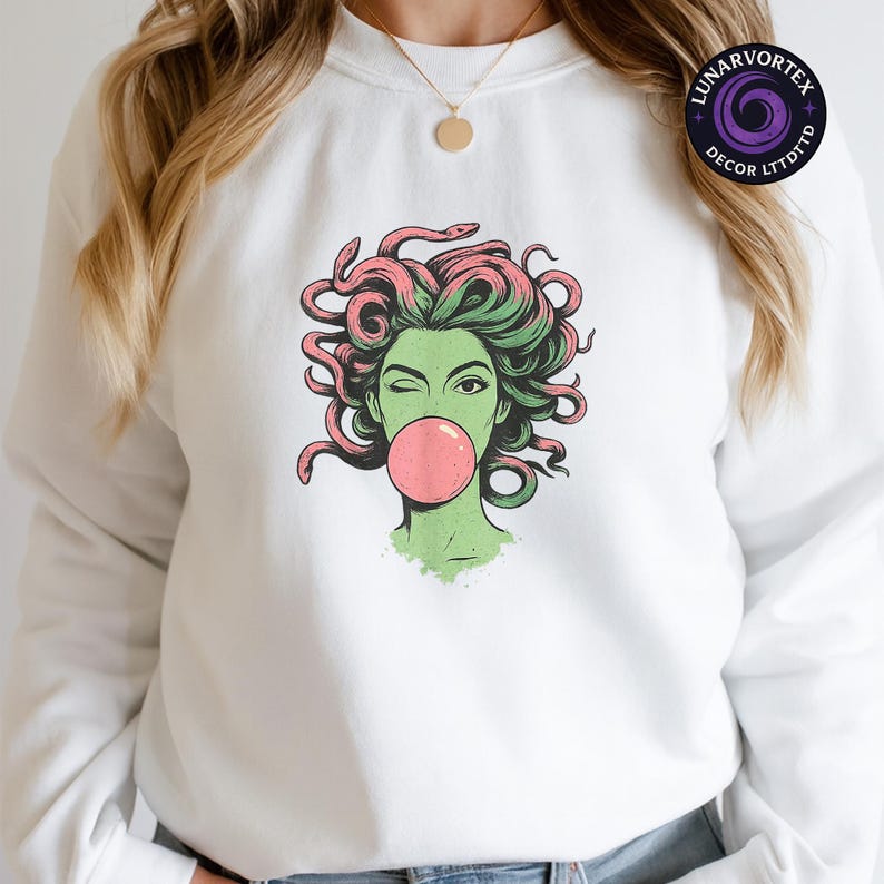 May include: White sweatshirt featuring a graphic of Medusa winking and blowing a bubblegum bubble. The Medusa illustration has green skin, green and pink hair, and is winking. The sweatshirt has a crew neck and long sleeves. The brand name "LUNARVORTEX DECOR LTDTTD" is printed on the upper right.