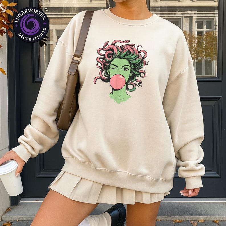 May include: Cream-colored sweatshirt with a graphic of a winking woman blowing a bubblegum bubble. The woman's skin is green, with pink and green hair. The sweatshirt is paired with a matching pleated skirt. A shoulder bag and a coffee cup are also visible.
