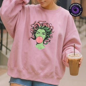 May include: A light pink sweatshirt featuring a graphic of a green-skinned woman with pink hair and snakes, blowing a pink bubble. The design is centered on the front of the sweatshirt.