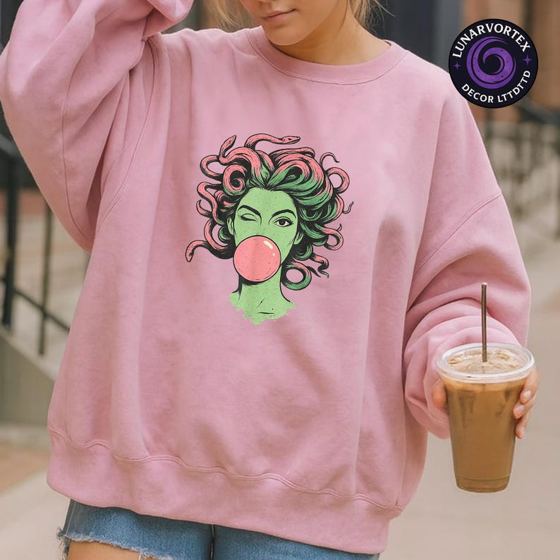 May include: A light pink sweatshirt featuring a graphic of a green-skinned woman with pink hair and snakes, blowing a pink bubble. The design is centered on the front of the sweatshirt.