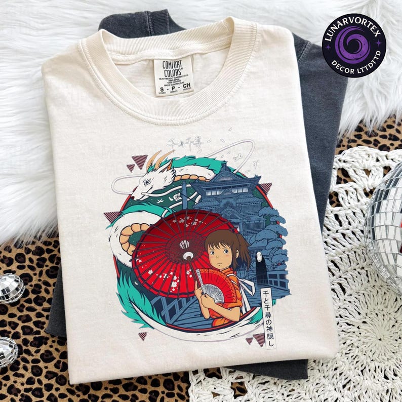 K&ouml;nnte beinhalten: Cremefarbenes T-Shirt mit einer farbenfrohen Grafik aus dem Zeichentrickfilm Spirited Away. Das Design umfasst einen Drachen, ein M&auml;dchen mit einem F&auml;cher und japanischen Text. Das Shirt ist von der Marke Comfort Colors.
