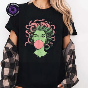 May include: Black t-shirt featuring a graphic of a winking woman with green skin, pink and green hair, and a pink bubblegum bubble. The design is a stylized illustration of Medusa. The shirt has a crew neck and short sleeves.