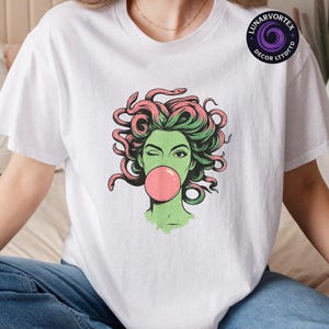 May include: White t-shirt featuring a graphic of a green-skinned woman with pink and green snake hair blowing a pink bubble. The woman is winking. The shirt has a round neck and short sleeves. The brand logo is in the upper right corner.