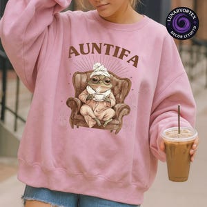 May include: Pink sweatshirt with the word "AUNTIFA" in brown text. The graphic features a frog in a robe, glasses, and a head wrap, seated in a brown armchair and holding a teacup. The sweatshirt is a solid pink color.