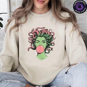 May include: Beige sweatshirt featuring a graphic of a green-skinned woman with pink and green hair, blowing a pink bubble. The design has a cartoonish style. The sweatshirt is a neutral color.