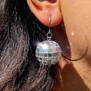 Op de afbeelding: Een bolvormige discobal-oorbel bestaande uit een veelvoud aan kleine, vierkante, reflecterende spiegeltjes. De zilverkleurige metalen haak gaat door de oorlel, waardoor de miniatuur discobal vrij aan een klein rond ringetje kan hangen.